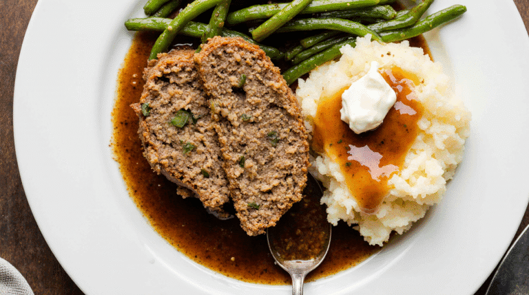 A delicious slice of homemade Cracker Barrel meatloaf recipe served with mashed potatoes, gravy, and green beans.